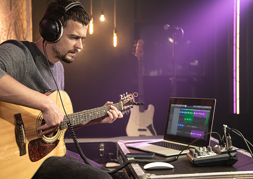 A male guitarist plays the guitar with headphones connected to a sound mixer. Digital waveform on laptop monitor, sound recording concept.