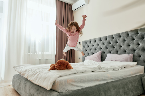 A cute little happy girl jumping down like a star on her teddybear barefoot on a bed in a huge bedroom. Happy childhood concept