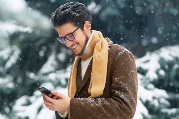 冬の公園で電話を持つ若い男。 ビジネスマンは電話を使用し、雪の森で冬の瞬間を楽しんでいます。降雪。 - snow glasses ストックフォトと画像