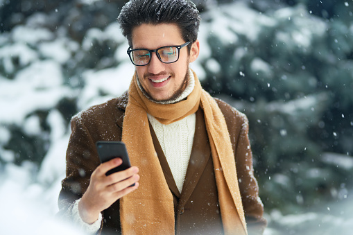 Young man with phone in winter park.  Businessman uses a phone and  enjoying winter moments in a snowy forest. Snowfall.