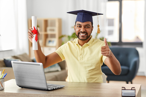 e-learning, education and people concept - happy smiling indian male graduate student with laptop computer and diploma showing thumbs up at home