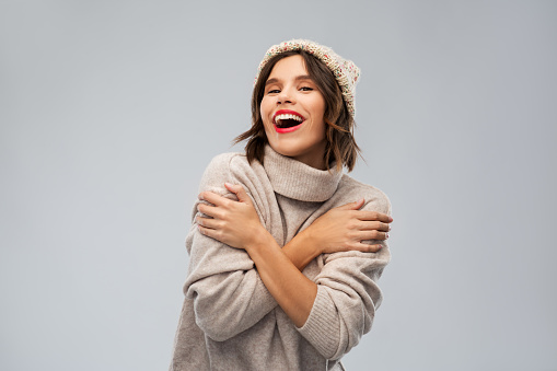 christmas, season and people concept - happy smiling young woman in knitted winter hat and sweater over grey background
