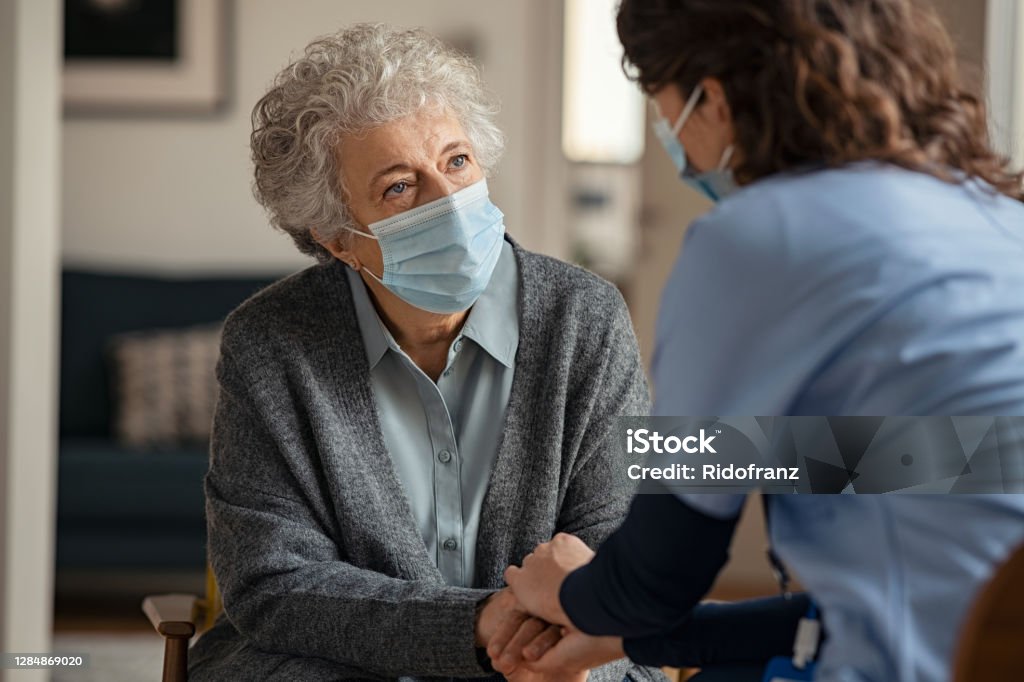 Female doctor consoling senior woman wearing face mask during home visit Elderly woman talking with a doctor while holding hands at home and wearing face protective mask. Worried senior woman talking to her general pratictioner visiting her at home during virus epidemic. Doctor explaining about precautionary measures during virus pandemic to old lady and takes care of her. Senior Adult Stock Photo