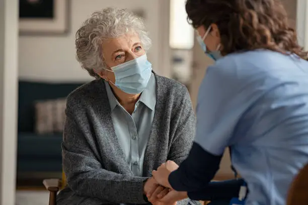 Elderly woman talking with a doctor while holding hands at home and wearing face protective mask. Worried senior woman talking to her general pratictioner visiting her at home during virus epidemic. Doctor explaining about precautionary measures during virus pandemic to old lady and takes care of her.