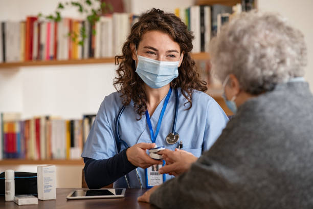 Doctor examining senior woman using oximeter at home Doctor wearing surgical mask while visiting a patient at home. Senior woman sitting with doctor while doing coronavirus test and screening using oximeter. Rear view of old woman with grey hair giving finger to doctor for oximeter analysis. oxygen photos stock pictures, royalty-free photos & images