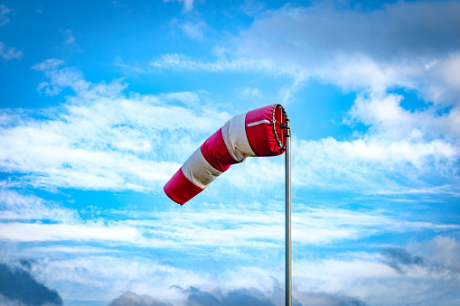 Red and white striped wind sock with clouds and blue sky in the background. The wind is coming from the right.