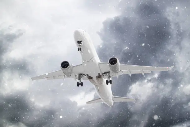 Photo of white passenger plane flies through falling snow against backdrop of beautiful ominous clouds on blue sky background in winter