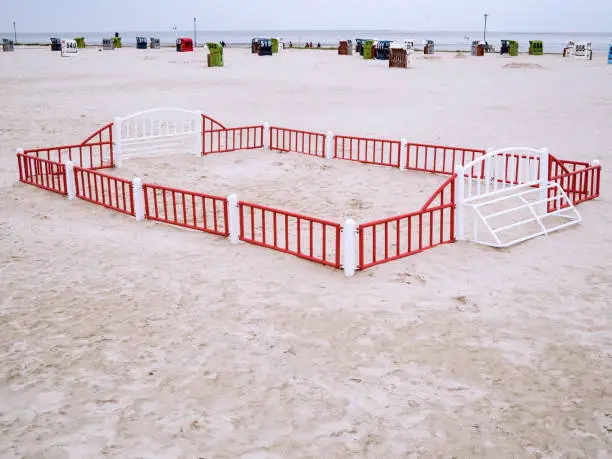German North Sea beach landscape with a small fenced-in beach ball playing field in the foreground and numbered wicker beach chairs far behind with a view over the Wadden Sea to the horizon when the sky is overcast.