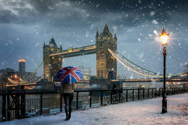 London winter concept with a tourist standing in front of the Tower Bridge with snowfall London winter concept with a tourist holding a british flag souvenir umbrella standing in front of the Tower Bridge during evening time with snowfall and cold temperature, United Kingdom winter wonderland london stock pictures, royalty-free photos & images