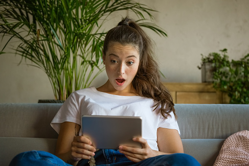 Shocked young beautiful woman wearing white t-shirt sitting on sofa in the living room and holding digital tablet, having video conference or watching movie.