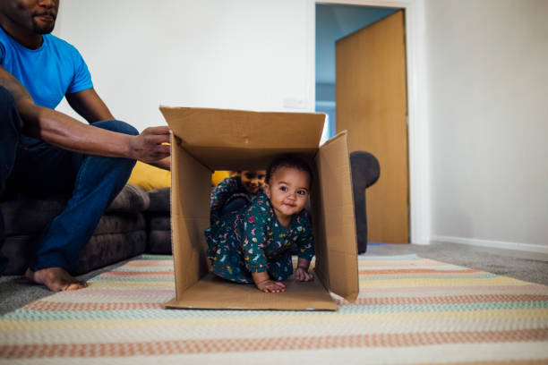 Playing at Home Mixed race baby girl crawling through a cardboard box in the sitting room at home, her father is holding the box up for her and her sister is in the background. obstacle course stock pictures, royalty-free photos & images