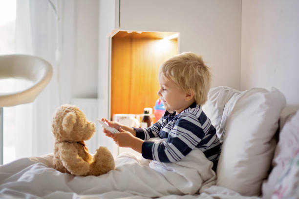 istock Little toddler blond child, playing with teddy bear in bed, while being sick, checking his temperature 1284893979