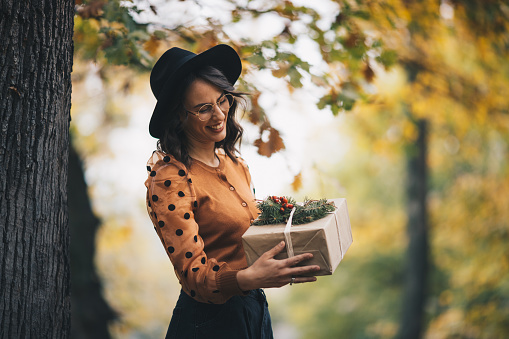 Smiling young hipster woman holding gift, leaning against a tree. She is astonished by wrapped present and decorated in branches and other natural materials.