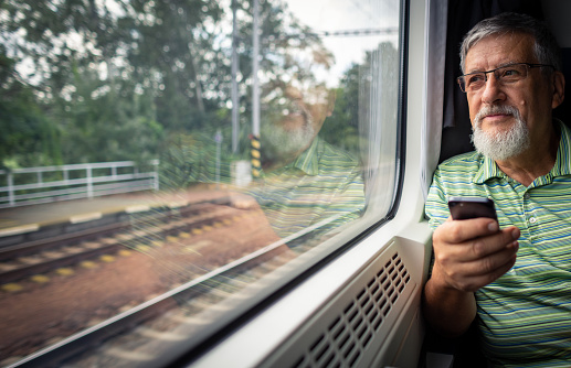 Senior man enjoying a train travel - leaving his car at home, he savours the time spent travelling, looks out of the window, has time to admire the landscape, use his smart phone to catch up with family