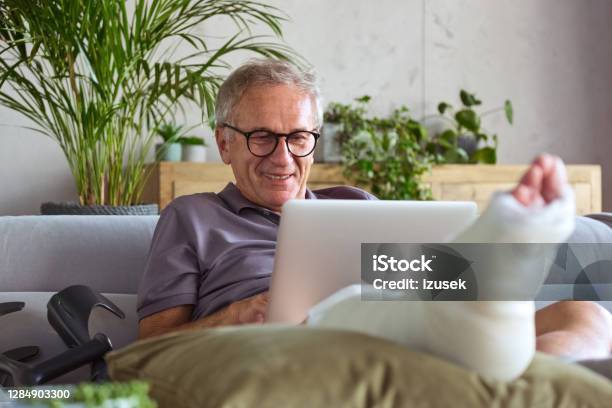 Cheerful Senior Man With Broken Leg At Home Stock Photo - Download Image Now - Broken Leg, Computer, Laptop