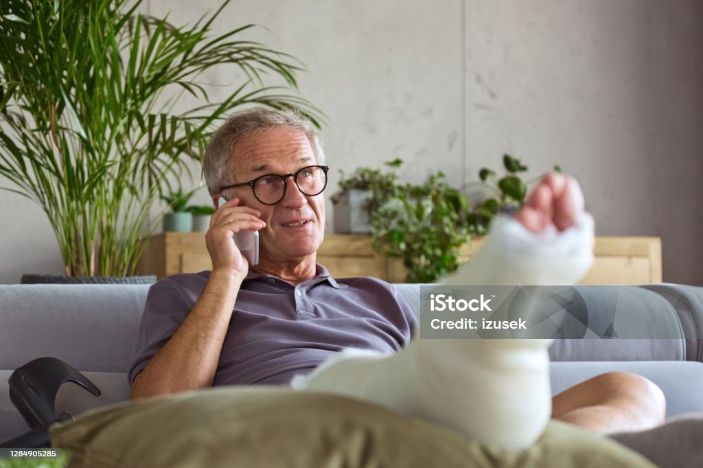 Senior man with broken leg at home Cheerful senior man with broken leg in plaster cast sitting on sofa at home and talking on smart phone. Recovery Stock Photo