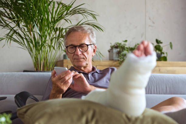 Senior man with broken leg at home Cheerful senior man with broken leg in plaster cast sitting on sofa at home and using smart phone. man sitting with a bandage on his leg stock pictures, royalty-free photos & images