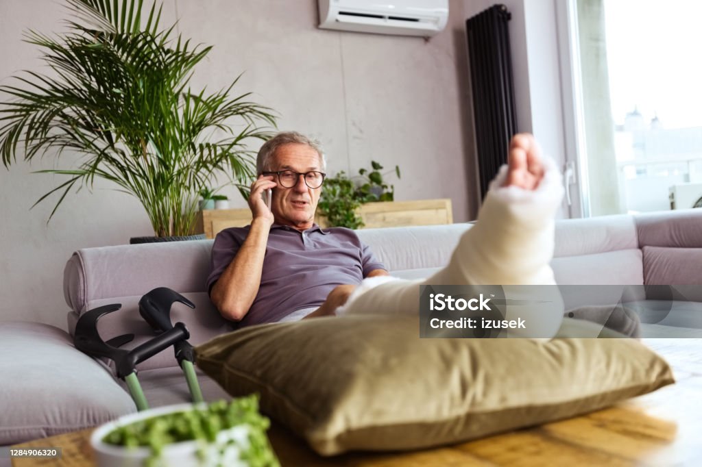 Senior man with broken leg at home Senior man with broken leg in plaster cast sitting on sofa at home and talking on smart phone. Crutch Stock Photo