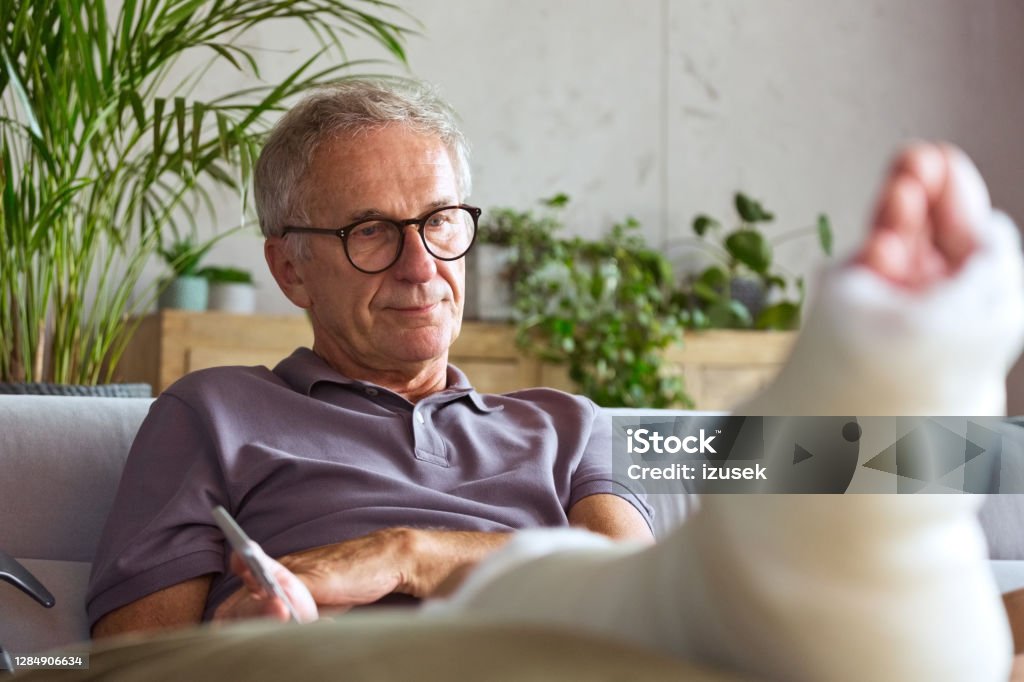 Worried senior man with broken leg at home Displeased senior man with broken leg in plaster cast sitting on sofa at home, looking worried. Orthopedic Cast Stock Photo
