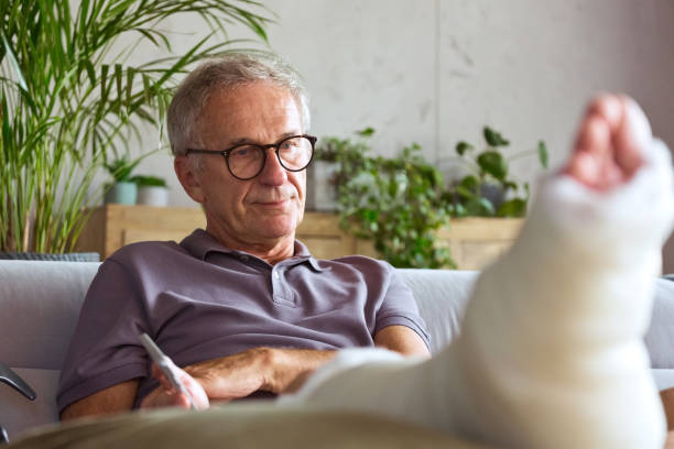 Worried senior man with broken leg at home Displeased senior man with broken leg in plaster cast sitting on sofa at home, looking worried. bone fracture stock pictures, royalty-free photos & images