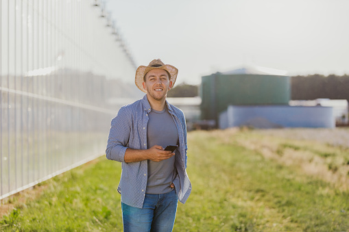 Waist up portrait of a smiling farmer in a hat holding his mobile phone near a greenhouse and looking at the camera