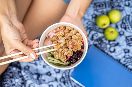 High angle of woman having picnic outdoor sitting on blue blanket with chopsticks eating takeaway healthy vegetarian poke bowl with tofu, fermented soybeans -tempeh, cucumber, rice, carrots, onion