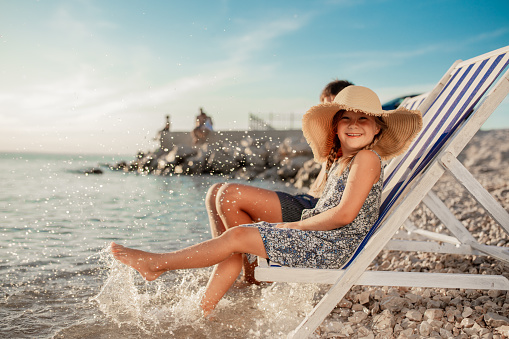 Smiling girl in a sun hat sitting on a deckchair on a beach in the summer, looking at the camera and splashing