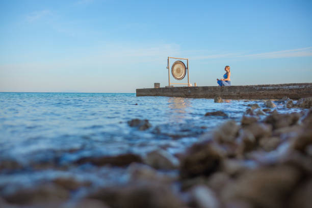 Woman sitting in lotus position doing yoga on pier with gong at dawn