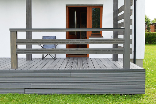 Part of white residential house with brown doors, windows and dark gray or anthracite wpc composite material terrace deck with wooden railings  in backyard outdoors.