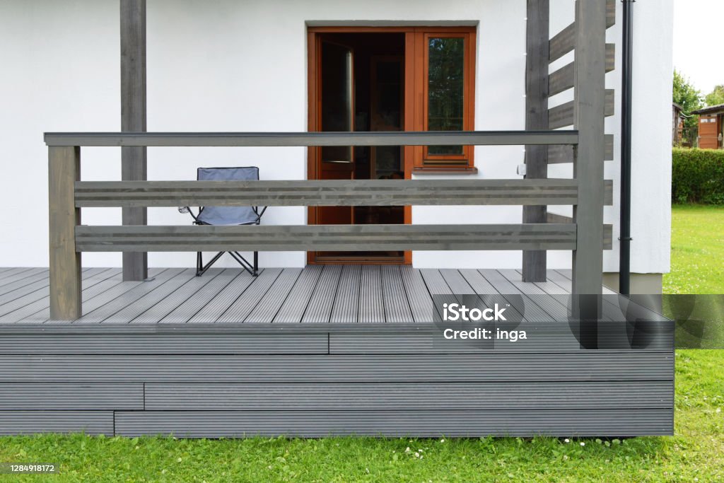 Part of white residential house with dark gray composite material terrace deck with wooden railings. Part of white residential house with brown doors, windows and dark gray or anthracite wpc composite material terrace deck with wooden railings  in backyard outdoors. Deck Stock Photo
