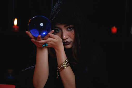 Portrait of Asian fortune teller in black hood holding blue crystal ball.