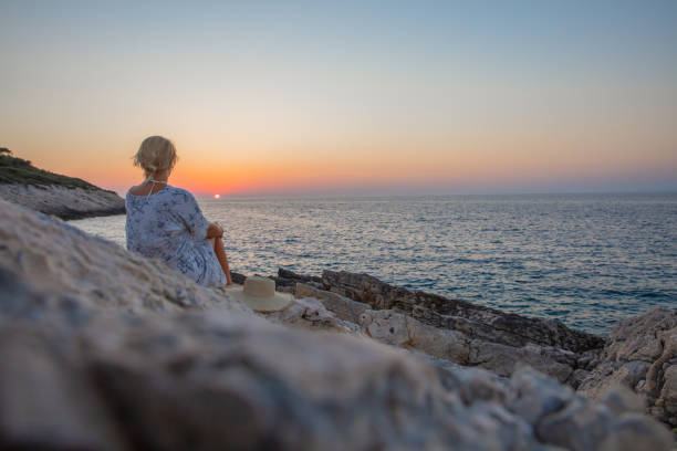 Blond woman sitting on beach and looking at romantic sunset sky over sea