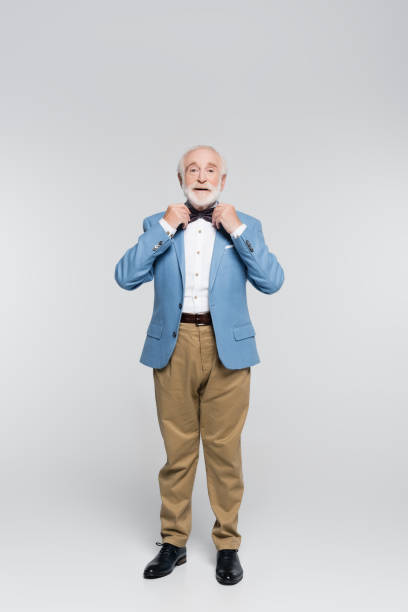 istock Smiling senior man in jacket adjusting bow tie on grey background 1284921338