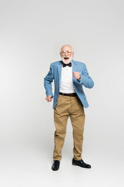 Cheerful senior man in jacket and bow tie dancing on grey background