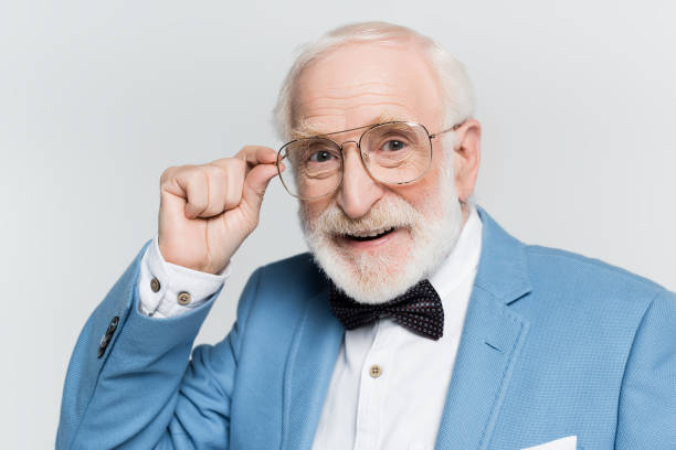 istock Positive senior man in blue jacket and bow tie touching eyeglasses isolated on grey 1284921415
