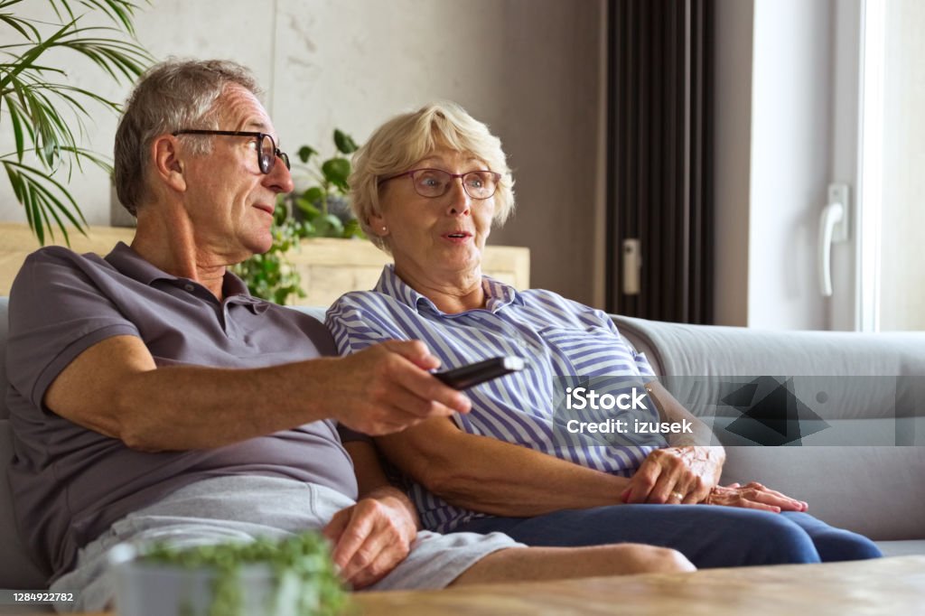 Senior couple watching tv at home Happy senior couple sitting on sofa in living room at home and watching tv. Man holding tv remote. Remote Control Stock Photo