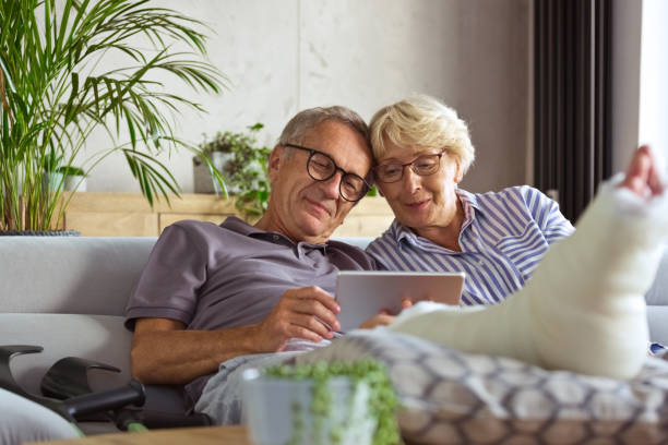 Senior man with broken leg at home and  his wife Happy senior couple sitting on sofa in living room at home, having video all. Senior man with broken leg. broken leg stock pictures, royalty-free photos & images