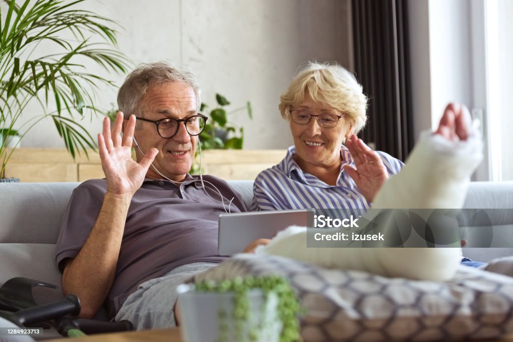 Senior man with broken leg at home and  his wife Cheerful senior couple sitting on sofa in living room at home, having video all. Senior man with broken leg. Senior Couple Stock Photo