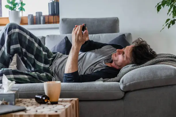 Photo of A sick man lying on the sofa and looking at his phone