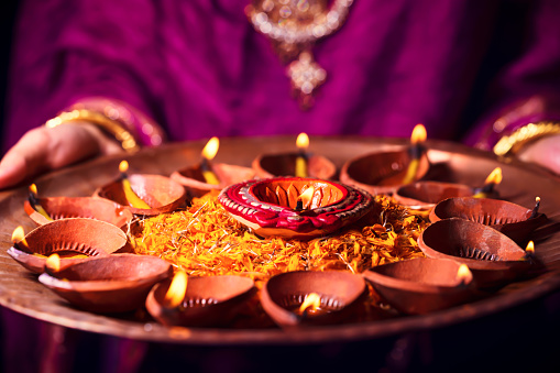 Happy Diwali Background. Indian woman or bride wearing traditional cloth and jewelry, holding puja thali full of lit burning diya or clay oil lamps, decorated with flowers.