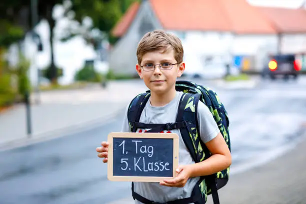 Happy little kid boy with glasses and backpack or satchel. Schoolkid on the way to middle or high school. Child outdoors on the street. Back to school. On desk First day fifth grade in German