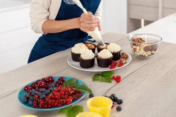 Photo of Female baker in blue apron  decorating tasty cupcake with creamy top at kitchen