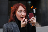 Redhead caucasian business woman looking at little pocket mirror and applying red lipstick after work