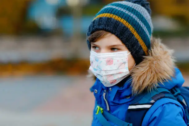 Little kid boy wearing medical mask on the way to school. Child backpack satchel. Schoolkid on cold autumn or winter day with warm clothes. Lockdown and quarantine time during corona pandemic disease.