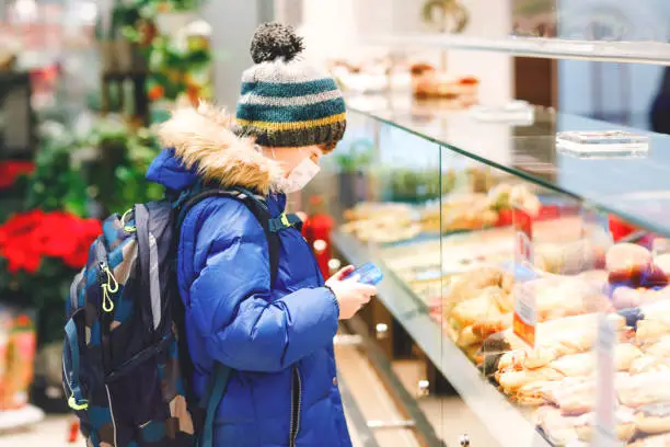 Kid boy wearing medical mask buy bread and pastry for school lunch in bakery. Child with backpack and winter clothes. Schoolkid during lockdown and quarantine time during corona pandemic disease.