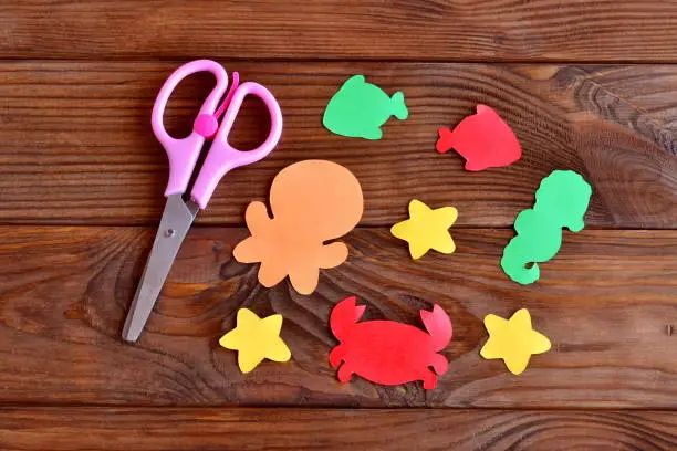 Photo of Marine animals cut from colored paper - octopus, fish, starfish, seahorse, crab. Kids sea animals arts and crafts. Scissors on wooden background. DIY summer activities for kids cheap craft ideas