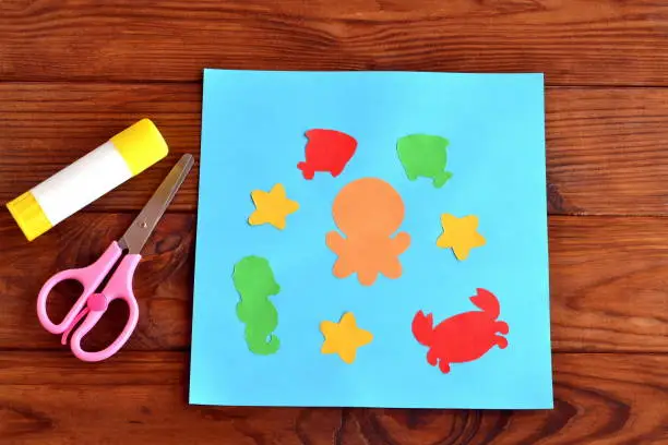 Photo of Paper card with sea animals and fishes. Creative ocean creatures crafts for kids. Paper octopus, fish, starfish, seahorse, crab project for children. Scissors, glue on a wooden table. Paper glue and cutting practice for kids