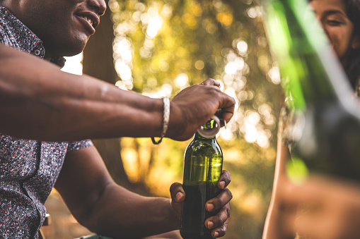 Low angle view of smiling man using a bottle opener to open a bottle of beer while enjoying a picnic with his friends.