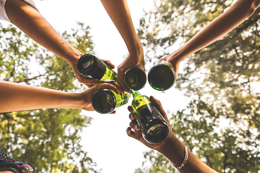 Directly below shot of five friends making celebratory toast with bottles of beer while hanging out outdoors together.