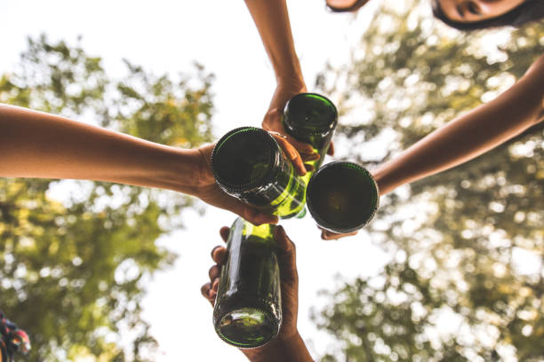 Directly below group of unrecognizable friends making celebratory toast with bottle of beer while on picnic in nature.